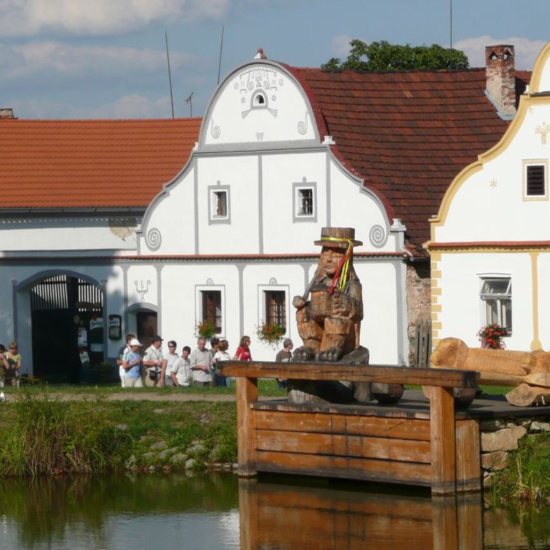 Malebný jihočeský barokní dům s vodníkem sedícím u rybníka v poklidné krajině.