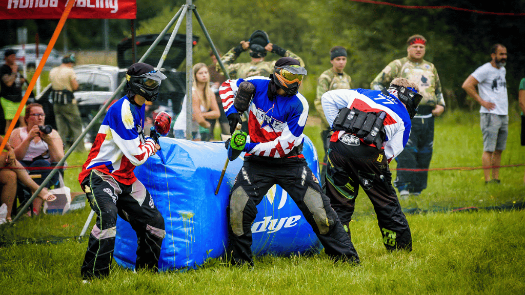Dvě osoby hrají paintball a skrývají se za překážkami.